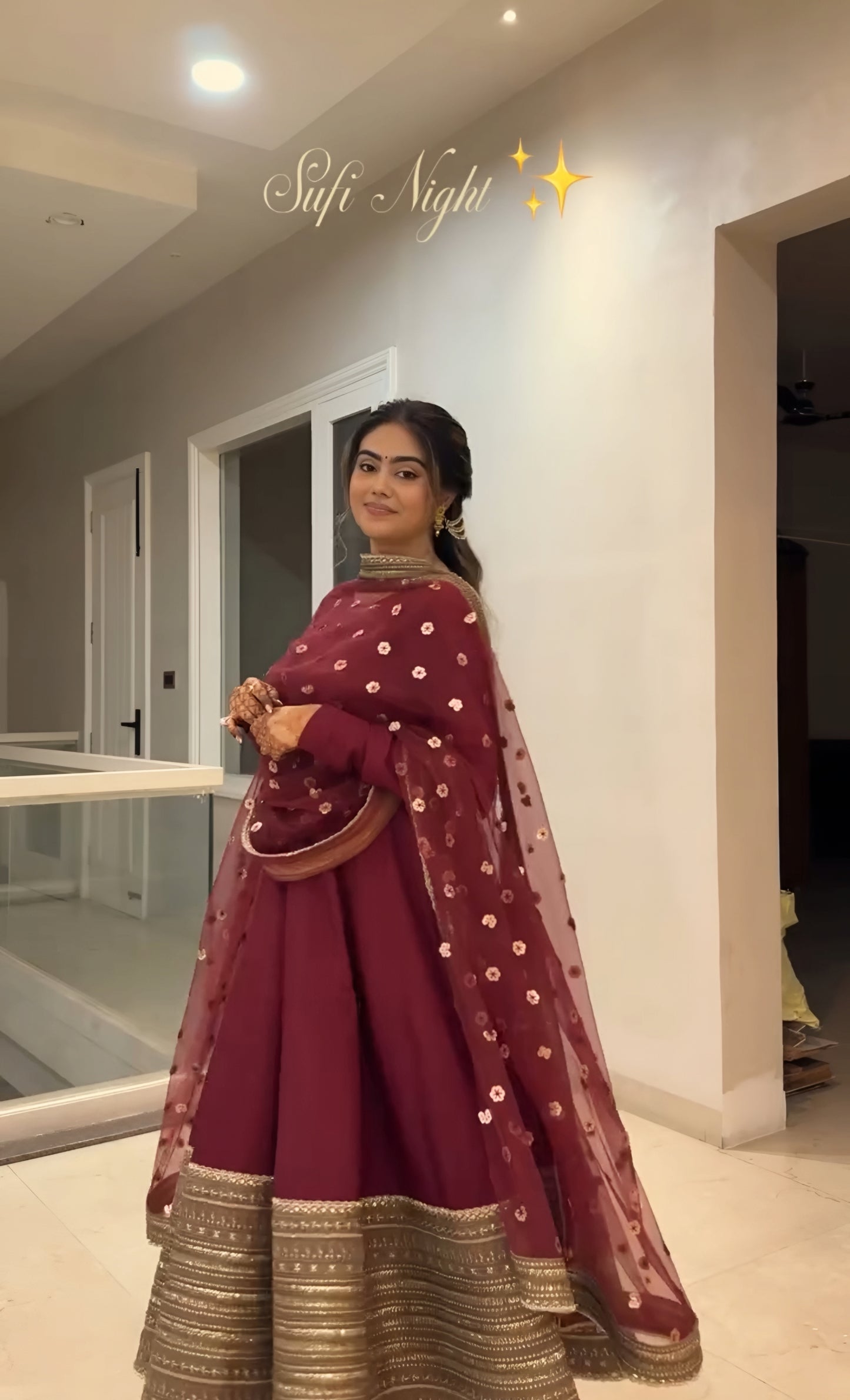 Woman in a maroon traditional outfit with gold details standing indoors.
