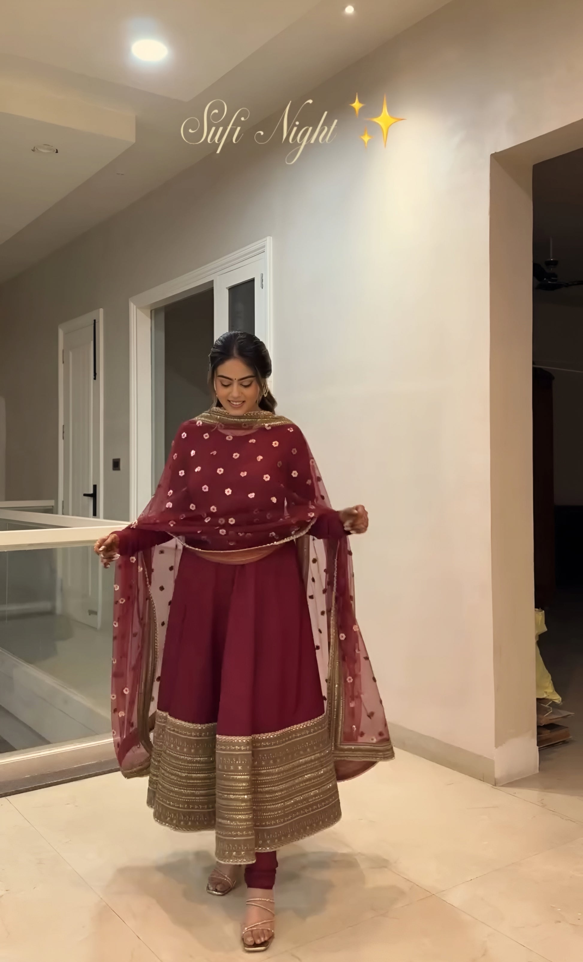 Woman in a traditional maroon and gold outfit standing in a room with neutral walls.