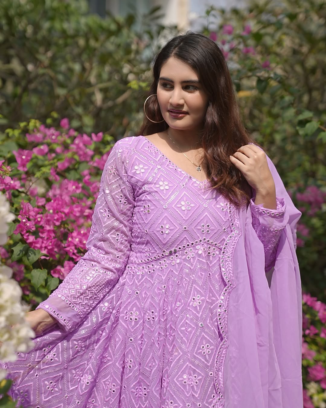 Woman in a purple embroidered outfit standing in a garden with pink flowers.