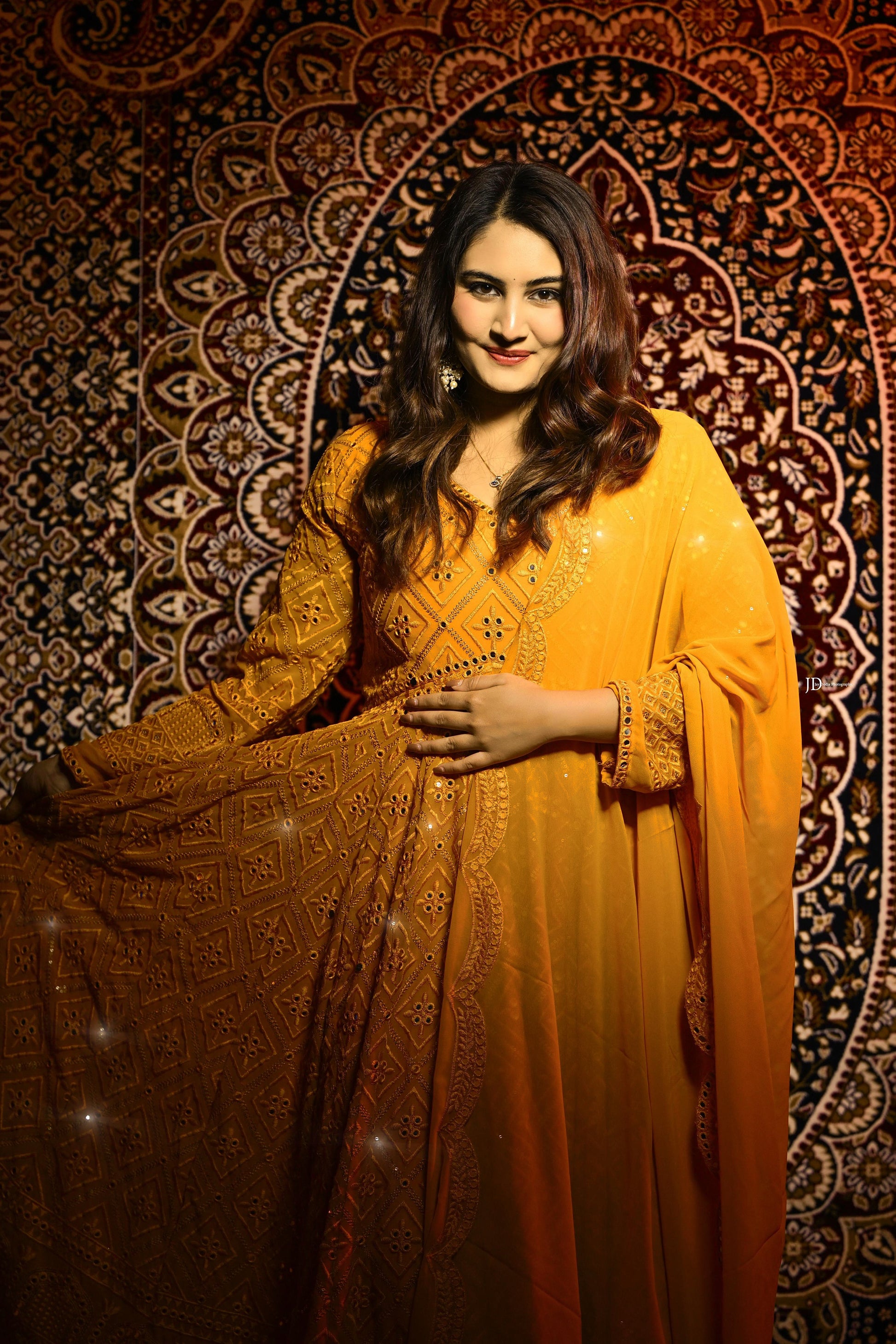 Woman in a yellow traditional outfit standing against an ornate patterned wall.